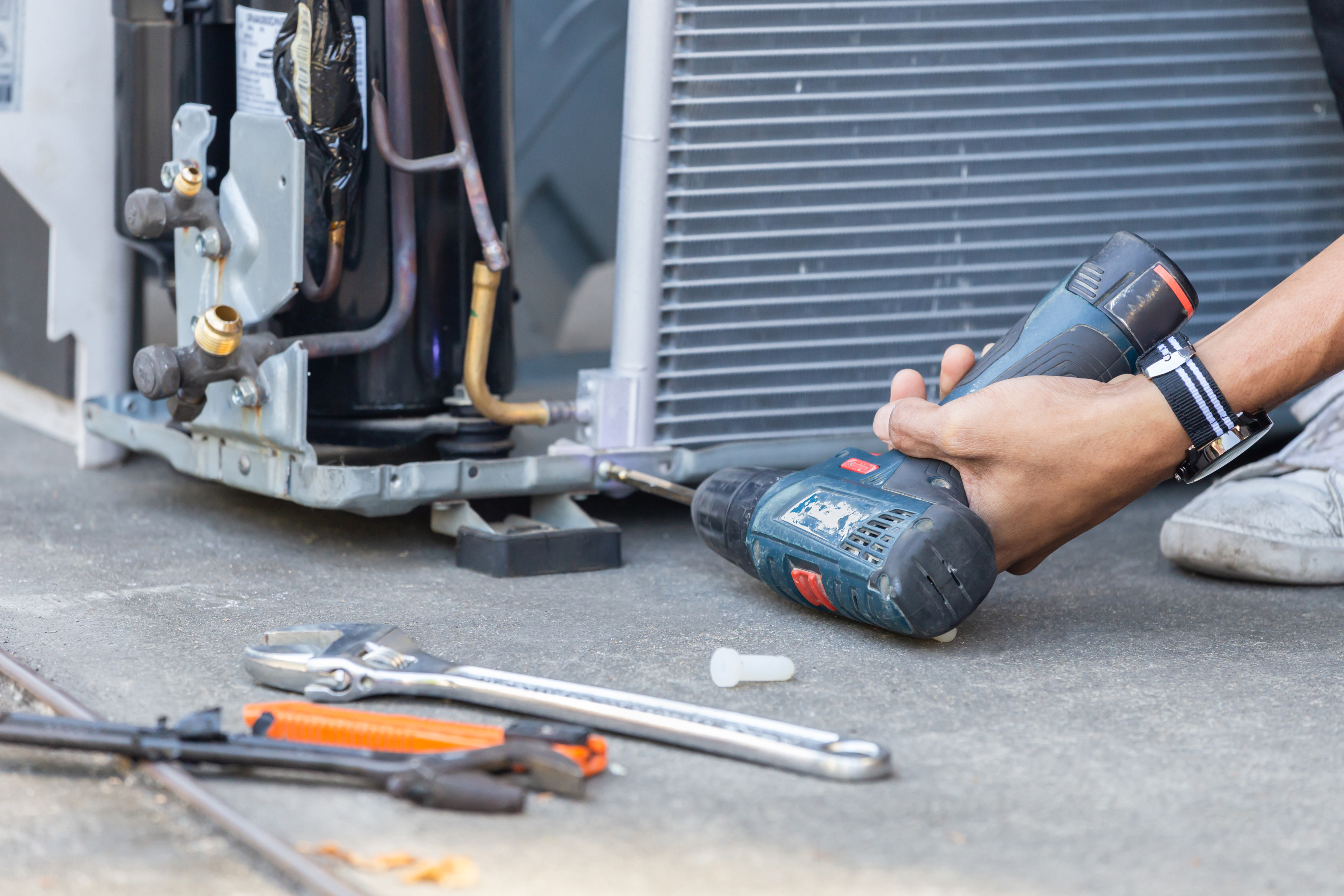 HVAC repair technician using a power drill to fix an air conditioning compressor unit, with tools scattered nearby for repair work.